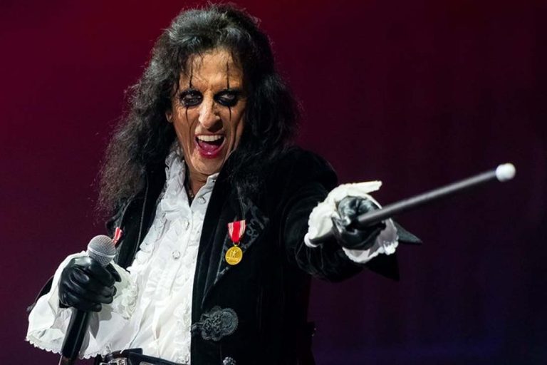 Alice Cooper performando no palco, usando roupas teatrais vitorianas, maquiagem marcante nos olhos, segurando um microfone e apontando uma bengala em direção ao público.