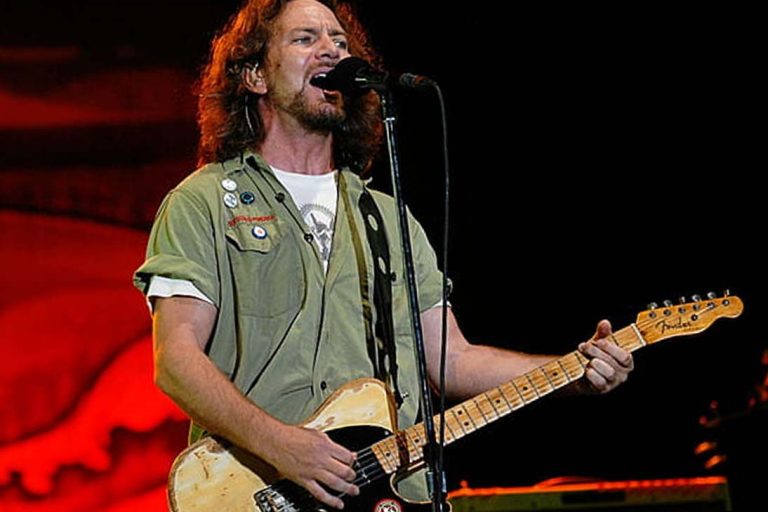 Músico cantando e tocando guitarra elétrica durante apresentação ao vivo.