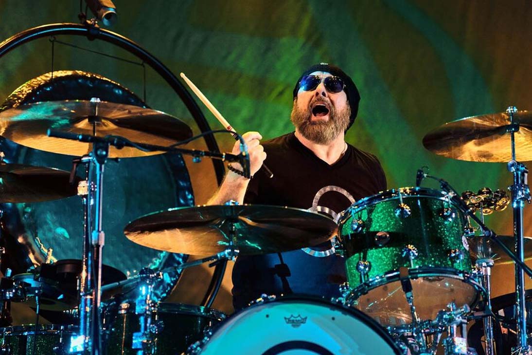 Jason Bonham tocando bateria em show tributo ao Led Zeppelin, com bateria verde metálica, usando touca e óculos escuros.