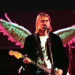 Kurt Cobain tocando guitarra em show com asas luminosas ao fundo.