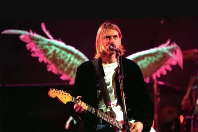 Kurt Cobain tocando guitarra em show com asas luminosas ao fundo.