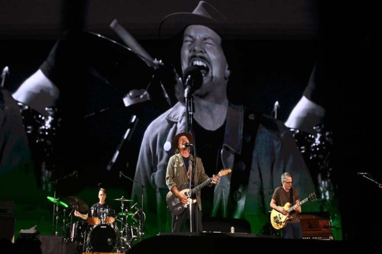 Integrantes da banda Pearl Jam se apresentando ao vivo com Eddie Vedder no centro, cantando com intensidade diante de uma grande projeção em preto e branco ao fundo.