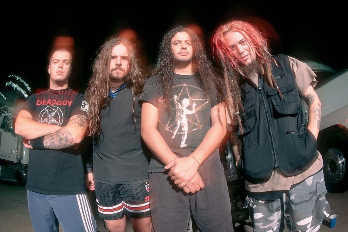 Integrantes da formação clássica da banda Sepultura posam em frente a caminhões, com roupas alternativas e visual característico do metal dos anos 90.
