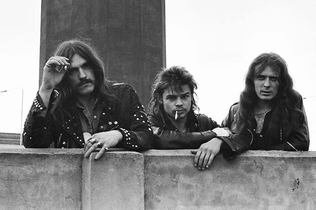 Foto da formação clássica do Motörhead apoiados em muro - Lemmy Kilmister, Eddie Clarke e Phil Taylor