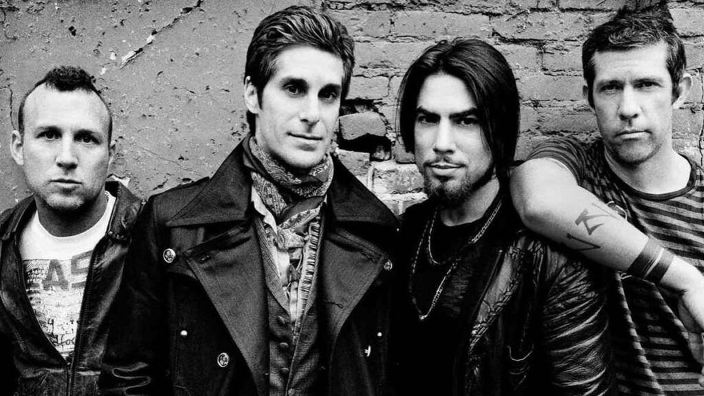 Foto em preto e branco dos integrantes da banda Jane's Addiction posando em frente a uma parede de tijolos.