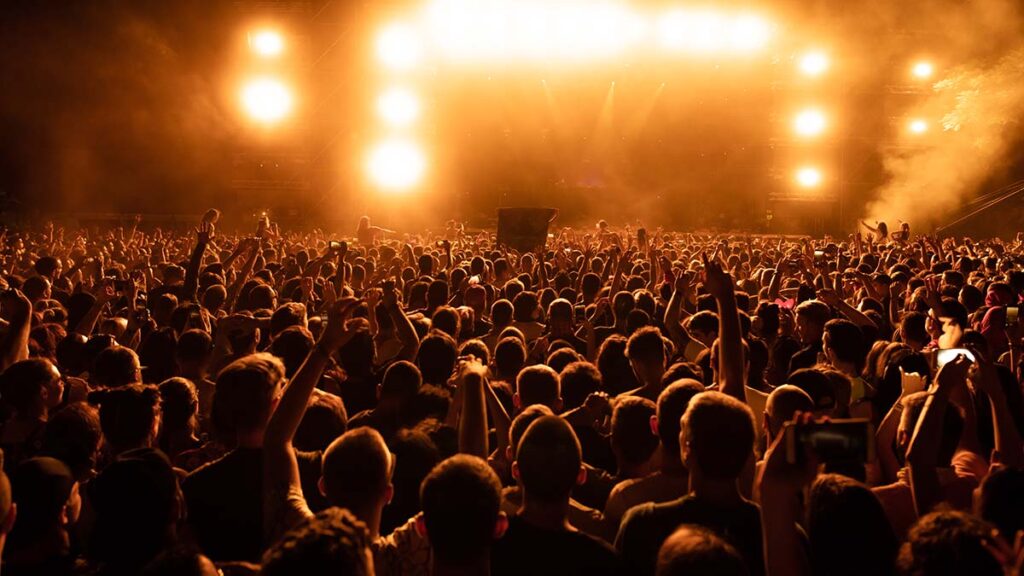 Multidão lotada em show de rock com mãos levantadas e celulares, palco iluminado com luzes alaranjadas vibrantes.