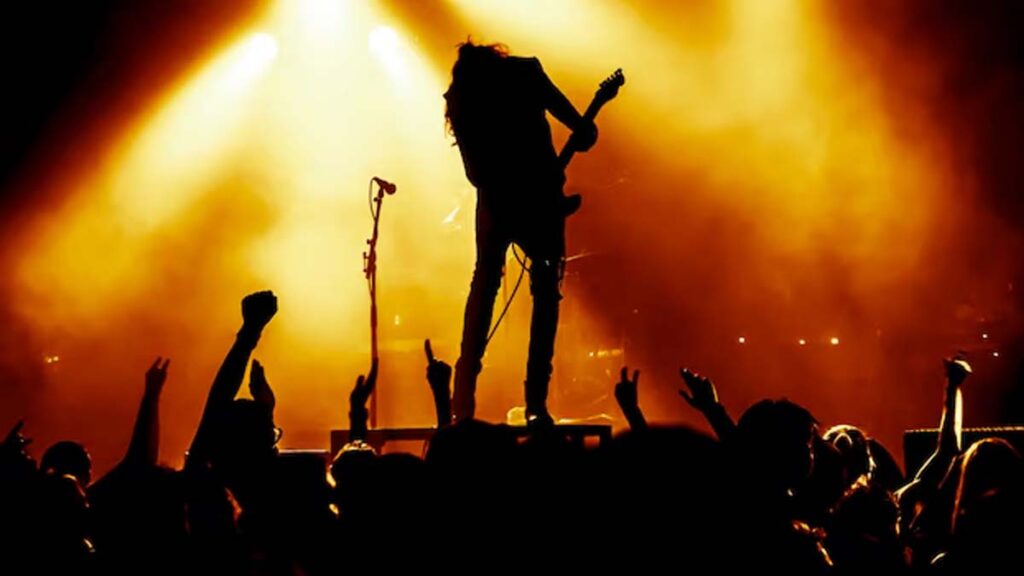 Silhueta de guitarrista em palco com luzes douradas tocando para multidão com braços erguidos em show de rock.