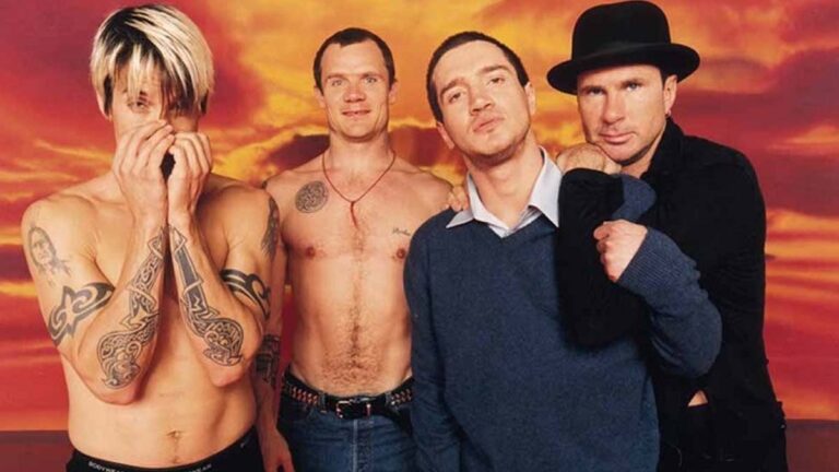 Integrantes da banda Red Hot Chili Peppers posando juntos em frente a um fundo colorido.